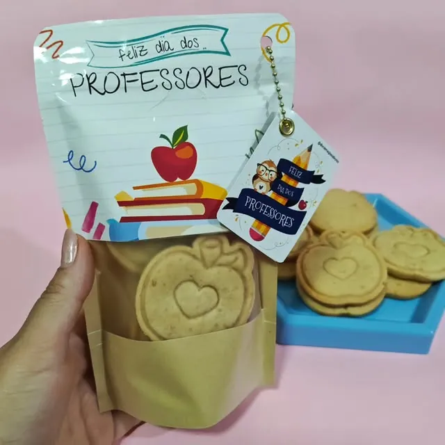 Pacote kraft com biscoitos carimbados de maçã e etiqueta Feliz Dia dos Professores, com biscoitos ao lado numa bandeja azul