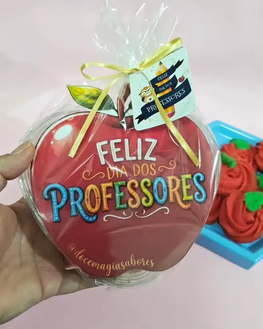 Mão segurando um biscoito grande de maçã com a frase Feliz Dia dos Professores, embalado em celofane com laço