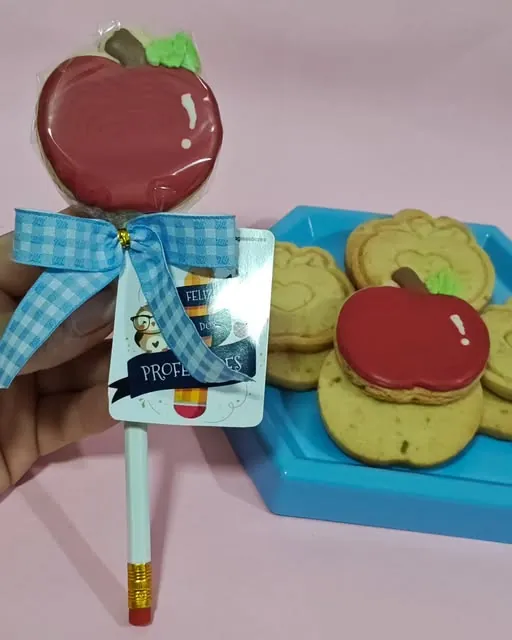 Biscoito de maçã vermelha decorado, preso a um lápis com laço xadrez azul, e outros biscoitos de maçã numa bandeja azul